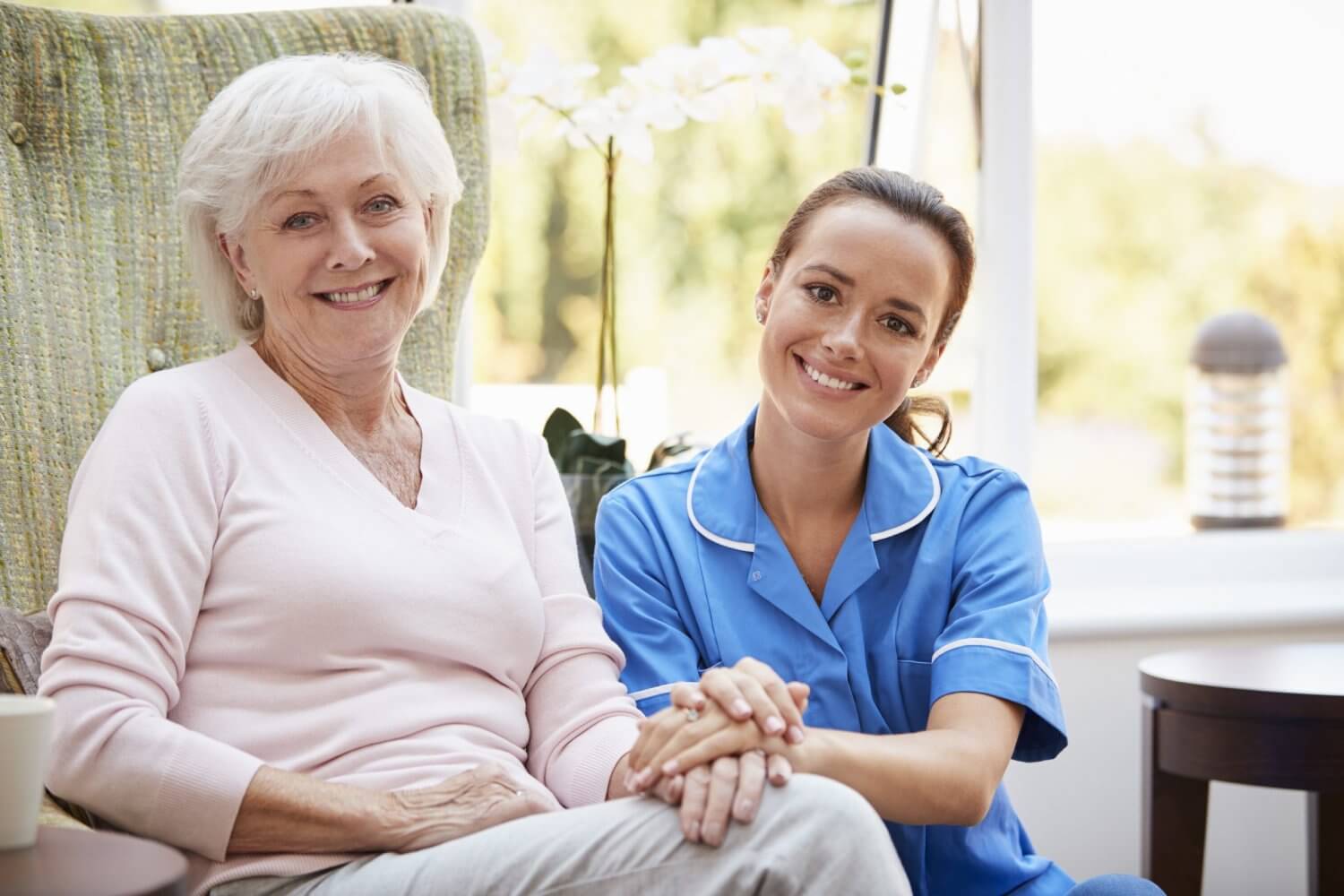 Portrait Of Senior Woman Sitting In Chair With Nurse In Retirement Home